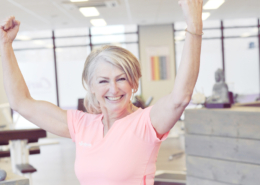 Sterke spieren krachttraining, 50+ vrouw traint met lichte gewichten in moderne fitnessruimte terwijl een coach haar begeleidt. Diverse vrouwen oefenen op hydraulische apparatuur in een warme, uitnodigende sfeer.