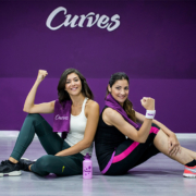 Vrouwvriendelijke fitnessruimte met hydraulische apparaten in cirkelopstelling waar twee 50+ vrouwen trainen onder begeleiding van een Curves-coach tijdens 30-minuten circuitprogramma.