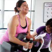 Vrouwen trainen samen op hydraulische apparatuur in een cirkelvormige Curves-fitnessruimte, begeleid door een coach in een warme, uitnodigende omgeving met paarse accenten.