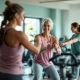 Vrouwen (50+) trainen samen in een uitnodigende fitnessruimte met hydraulische apparatuur in cirkelopstelling. Een coach begeleidt vriendelijk een deelnemer in een warme, paars-groene omgeving.