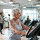 Twee 50+ vrouwen trainen met lichte gewichten in een warme fitnesszaal met paarse accenten, terwijl een coach vriendelijk begeleiding biedt in een veilige, ondersteunende omgeving.