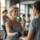 50+ vrouw traint met lichte gewichten in moderne fitnessruimte terwijl een coach haar begeleidt. Diverse vrouwen oefenen op hydraulische apparatuur in een warme, uitnodigende sfeer.
