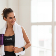 Vrouw van middelbare leeftijd traint zelfverzekerd op hydraulisch apparaat in lichte Curves fitnessruimte, terwijl coach andere vrouwen begeleidt in circuittraining.