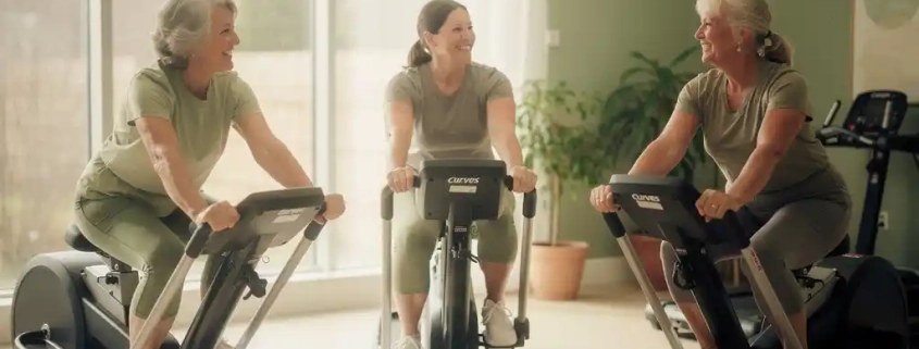 Drie vrouwen boven de 50 trainen samen op Curves hydraulische fitnessapparatuur in een lichte sportschool met natuurlijk licht
