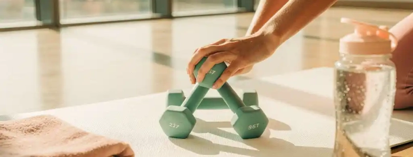 Vrouwenhanden houden drie kleine halters van verschillende gewichten in driehoekpatroon op witte yogamat in fitnessstudio