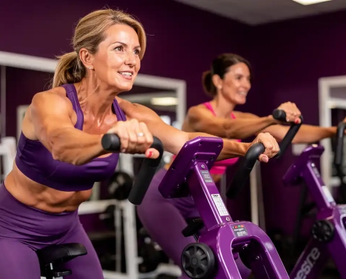Twee vrouwen van middelbare leeftijd trainen samen op paarse hydraulische fitnessmachines in een vrouwengerichte sportschool.