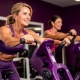 Twee vrouwen van middelbare leeftijd trainen samen op paarse hydraulische fitnessmachines in een vrouwengerichte sportschool.