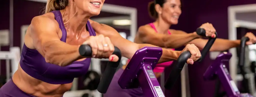 Twee vrouwen van middelbare leeftijd trainen samen op paarse hydraulische fitnessmachines in een vrouwengerichte sportschool.