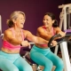 Twee vrouwen van middelbare leeftijd lachen en sporten samen op hydraulische fitnessmachines in een vrouwenfitnessclub met paarse muren.