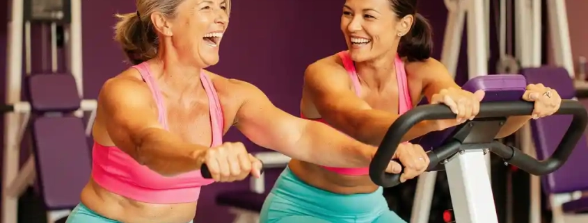 Twee vrouwen van middelbare leeftijd lachen en sporten samen op hydraulische fitnessmachines in een vrouwenfitnessclub met paarse muren.