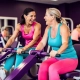 Twee vrouwen van middelbare leeftijd lachen samen tijdens krachttraining op paarse hydraulische machines in een levendige damesfitnesclub.