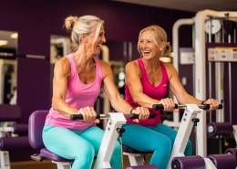 Twee vrouwen van middelbare leeftijd trainen lachend op hydraulische weerstandsmachines in een paars ingerichte damesfitnessclub.