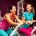 Twee vrouwen van middelbare leeftijd lachen samen tijdens een work-out op hydraulische fitnessapparaten in een paars ingerichte damesgymnastieksaal.