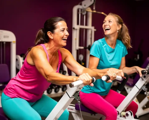 Twee vrouwen van middelbare leeftijd lachen samen tijdens een work-out op hydraulische fitnessapparaten in een paars ingerichte damesgymnastieksaal.