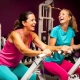 Twee vrouwen van middelbare leeftijd lachen samen tijdens een work-out op hydraulische fitnessapparaten in een paars ingerichte damesgymnastieksaal.