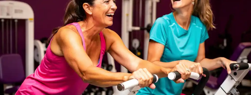 Twee vrouwen van middelbare leeftijd lachen samen tijdens een work-out op hydraulische fitnessapparaten in een paars ingerichte damesgymnastieksaal.