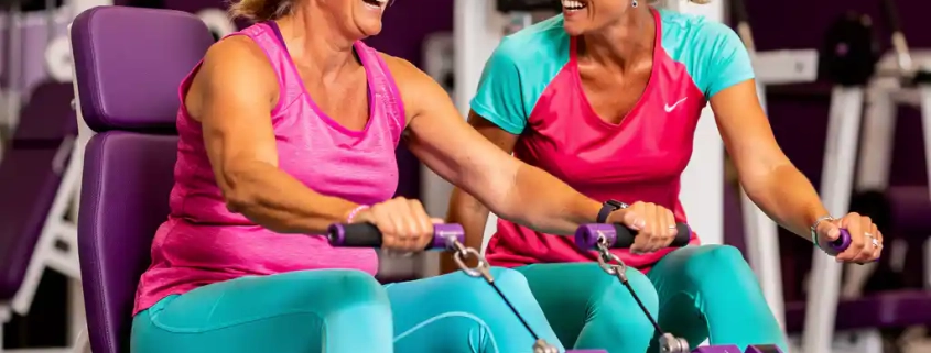 Twee vrouwen van middelbare leeftijd lachen samen tijdens krachttraining op paarse hydraulische machines in een lichte damesfitnessstudio.