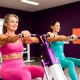 Twee vrouwen van middelbare leeftijd trainen lachend op paarse hydraulische fitnessmachines in een levendige damesfitnessclub met paarse muren.