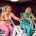 Twee vrouwen van middelbare leeftijd lachen tijdens krachttraining op hydraulische fitnessmachines in een vrouwenfitnessclub met paarse muren.