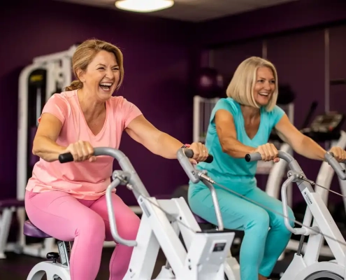 Twee vrouwen van middelbare leeftijd lachen tijdens krachttraining op hydraulische fitnessmachines in een vrouwenfitnessclub met paarse muren.