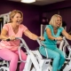 Twee vrouwen van middelbare leeftijd lachen tijdens krachttraining op hydraulische fitnessmachines in een vrouwenfitnessclub met paarse muren.