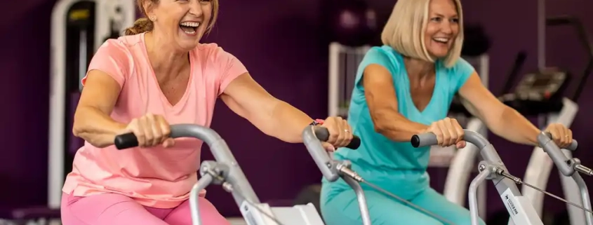 Twee vrouwen van middelbare leeftijd lachen tijdens krachttraining op hydraulische fitnessmachines in een vrouwenfitnessclub met paarse muren.