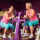 Twee vrouwen van middelbare leeftijd lachen samen tijdens krachttraining op paarse hydraulische machines in een vrouwenfitnessclub.