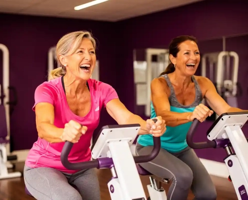 Twee vrouwen van middelbare leeftijd trainen lachend op hydraulische fitnessmachines in een paars ingerichte damesfitnessstudio.