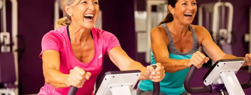 Twee vrouwen van middelbare leeftijd trainen lachend op hydraulische fitnessmachines in een paars ingerichte damesfitnessstudio.