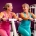 Twee vrouwen van middelbare leeftijd sporten samen op hydraulische fitnessmachines in een paarse damesgymnastiek, lachend en elkaar aanmoedigend.