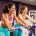 Twee vrouwen van middelbare leeftijd lachen samen op hydraulische weerstandsmachines in een felpaarse damesfitnessclub.