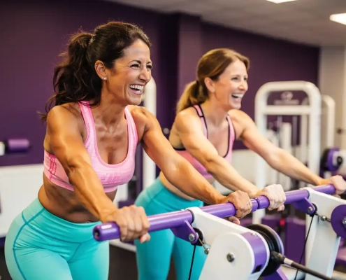 Twee vrouwen van middelbare leeftijd lachen samen op hydraulische weerstandsmachines in een felpaarse damesfitnessclub.