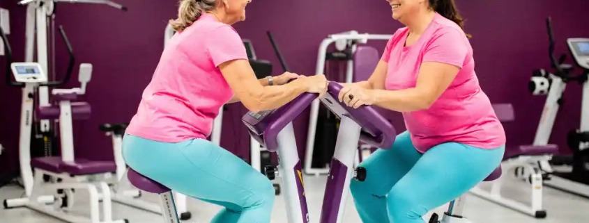 Twee vrouwen van middelbare leeftijd trainen enthousiast op hydraulische fitnessapparaten in een moderne damesgymnastiek met paarse muren.
