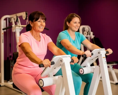 Twee vrouwen van middelbare leeftijd trainen samen op witte hydraulische fitnessapparaten in een levendige damesgymnastiek met paarse muren.
