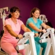 Twee vrouwen van middelbare leeftijd trainen samen op witte hydraulische fitnessapparaten in een levendige damesgymnastiek met paarse muren.