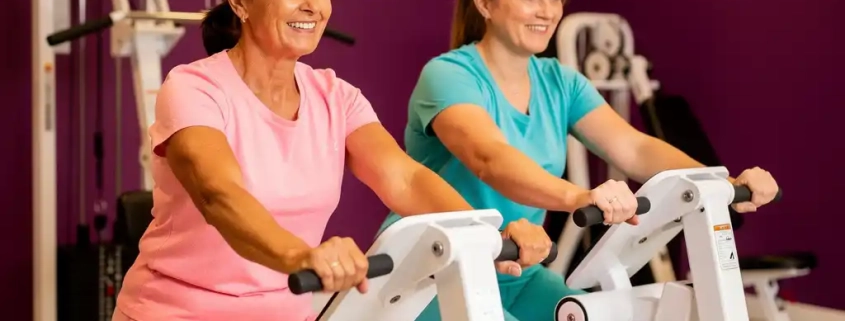 Twee vrouwen van middelbare leeftijd trainen samen op witte hydraulische fitnessapparaten in een levendige damesgymnastiek met paarse muren.