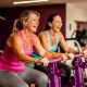 Twee vrouwen lachen samen tijdens een krachttraining op paarse hydraulische fitnessapparaten in een vrouwenfitnessclub.