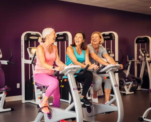 Drie vrouwen van middelbare leeftijd lachen samen tijdens een training op hydraulische fitnessapparaten in een paarse vrouwengymnastiekzaal.