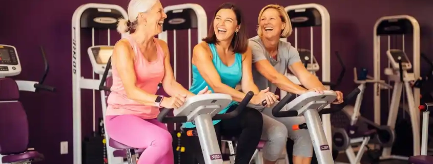 Drie vrouwen van middelbare leeftijd lachen samen tijdens een training op hydraulische fitnessapparaten in een paarse vrouwengymnastiekzaal.