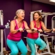 Twee vrouwen van middelbare leeftijd lachen samen tijdens krachttraining op hydraulische fitnessapparaten in een levendige vrouwengymnastiekzaal met paarse muren.