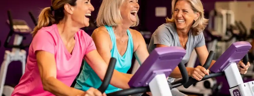 Drie lachende vrouwen tussen 40 en 60 jaar trainen samen op hydraulische fitnessapparaten in een levendige damesgymnastiek met paarse muren.