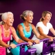 Drie vrouwen van 50-60 jaar lachen samen op paarse hydraulische fitnessmachines in een vrouwengymnastieklokaal met paarse muren.