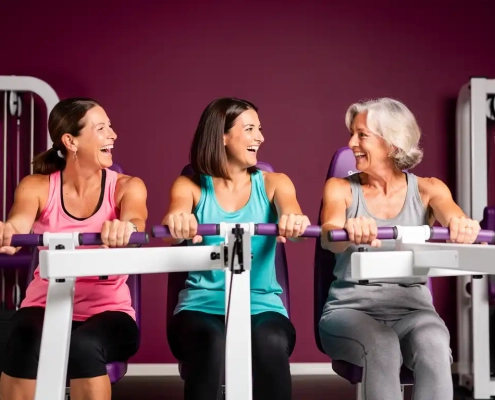 Drie vrouwen van middelbare leeftijd lachen samen tijdens een krachttraining op hydraulische fitnessapparaten in een paarse damesgymnastiek.