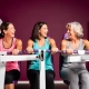 Drie vrouwen van middelbare leeftijd lachen samen tijdens een krachttraining op hydraulische fitnessapparaten in een paarse damesgymnastiek.