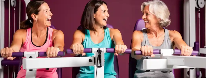 Drie vrouwen van middelbare leeftijd lachen samen tijdens een krachttraining op hydraulische fitnessapparaten in een paarse damesgymnastiek.