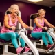 Twee vrouwen van 50-60 jaar lachen en sporten samen op hydraulische fitnessapparaten in een paarse damesgymnastiekzaal.
