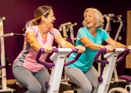 Twee vrouwen van in de vijftig lachen samen tijdens een training op hydraulische fitnessapparaten in een vrouwengym met paarse muren.