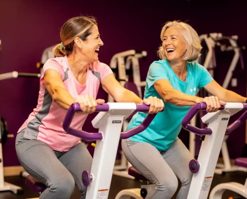 Twee vrouwen van in de vijftig lachen samen tijdens een training op hydraulische fitnessapparaten in een vrouwengym met paarse muren.