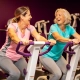 Twee vrouwen van in de vijftig lachen samen tijdens een training op hydraulische fitnessapparaten in een vrouwengym met paarse muren.