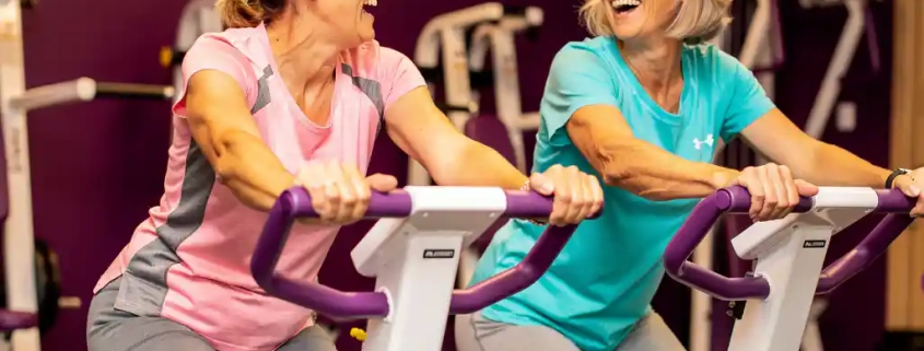 Twee vrouwen van in de vijftig lachen samen tijdens een training op hydraulische fitnessapparaten in een vrouwengym met paarse muren.